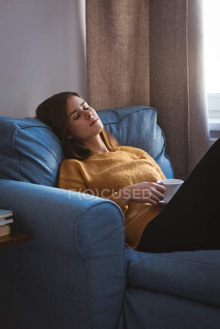 Jovem relaxante no sofá segurando uma xícara de café na sala de estar em casa — Fotografia de Stock