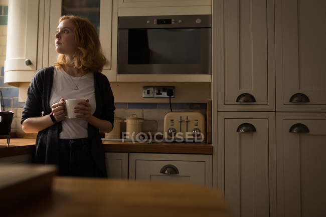 Jovem mulher segurando caneca e de pé na cozinha em casa — Fotografia de Stock