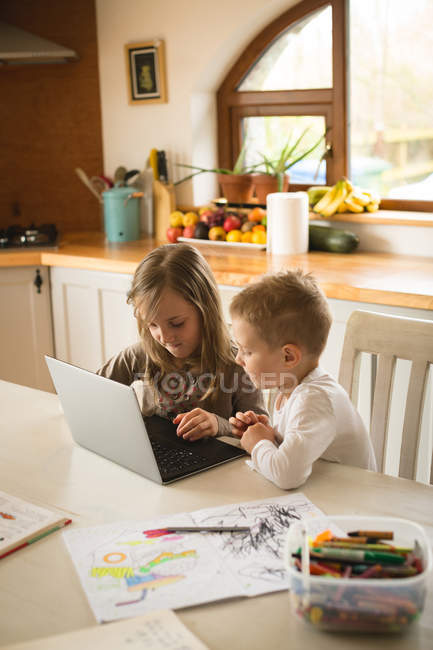 Los niños que usan el ordenador portátil juntos en la cocina en casa - foto de stock