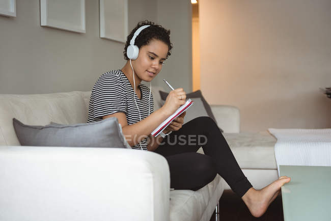 Woman listening to music while writing in notebook at home — Stock Photo