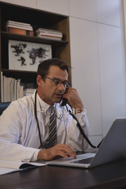 Доктор розмовляє по стаціонарній лінії під час використання ноутбука в офісі . — стокове фото