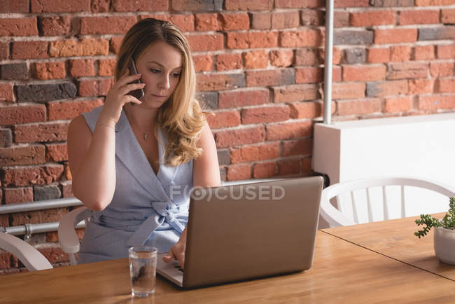 Female executive talking on mobile phone while using laptop in creative office — Stock Photo