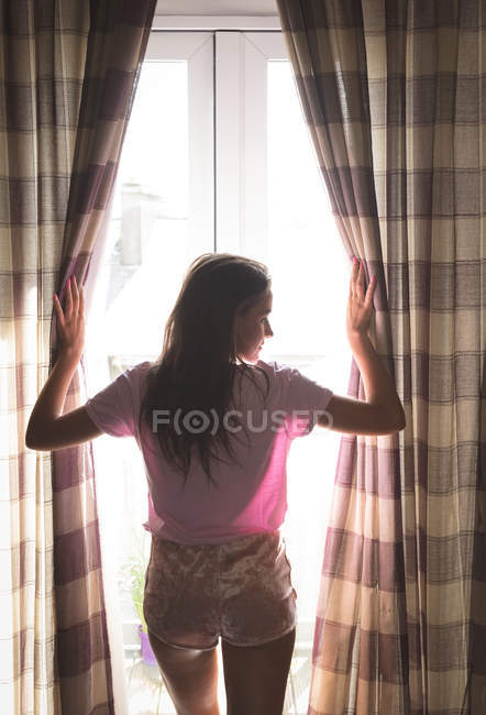 Visão traseira da mulher abrindo cortinas de janela em casa . — Fotografia de Stock
