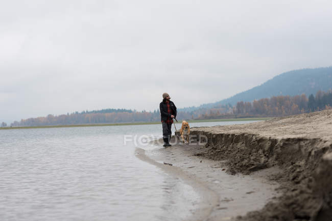 Homem e seu cão de estimação andando na margem do rio no dia nebuloso — Fotografia de Stock