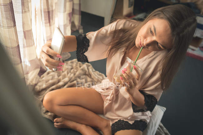 Mujer en ropa de dormir tomando selfie con teléfono móvil mientras toma café en casa . - foto de stock