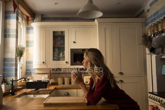 Mujer joven apoyada en el mostrador de la cocina tomando café en la cocina - foto de stock