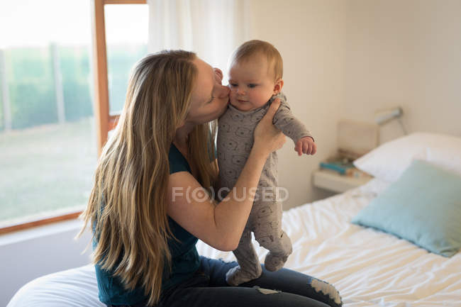 Mother carrying and kissing baby son on bed at home. — Stock Photo