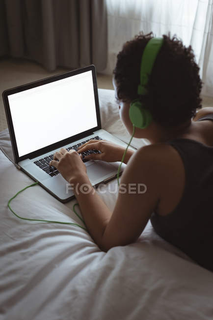 Frau benutzt Laptop, während sie Musik auf dem Bett im Schlafzimmer hört — Stockfoto