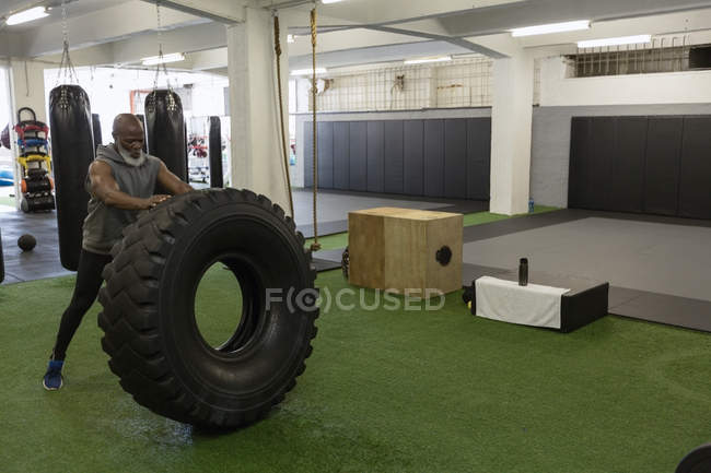 Determined senior man doing cross fit tire flip in fitness studio. — Stock Photo