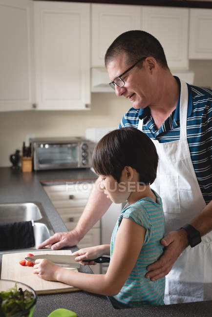 Padre atento ayudando a su hijo a cortar verduras en la cocina - foto de stock