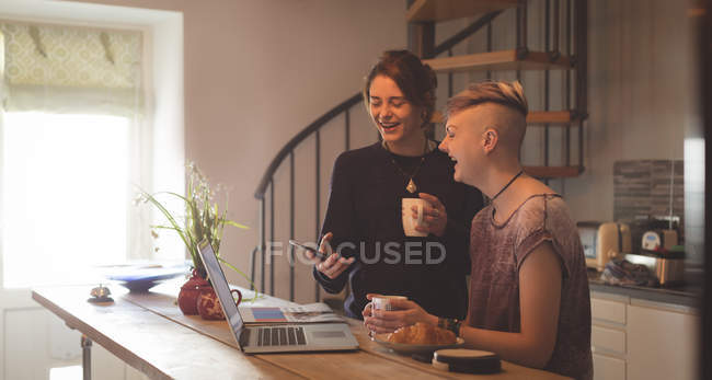 Щаслива лесбіянка пара використовує мобільний телефон і ноутбук, маючи каву вдома . — стокове фото