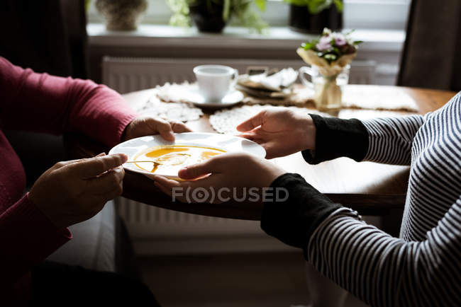 Partie médiane de la grand-mère recevant une assiette de soupe de la petite-fille dans le salon — Photo de stock
