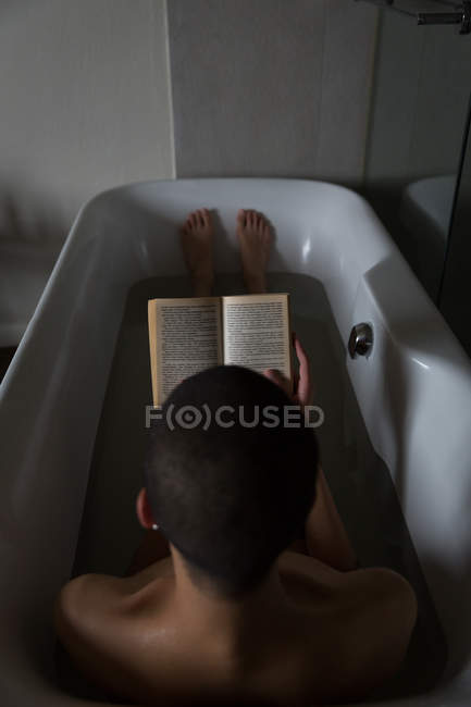 Jeune homme lisant le livre tout en étant couché dans la baignoire à la salle de bain — Photo de stock