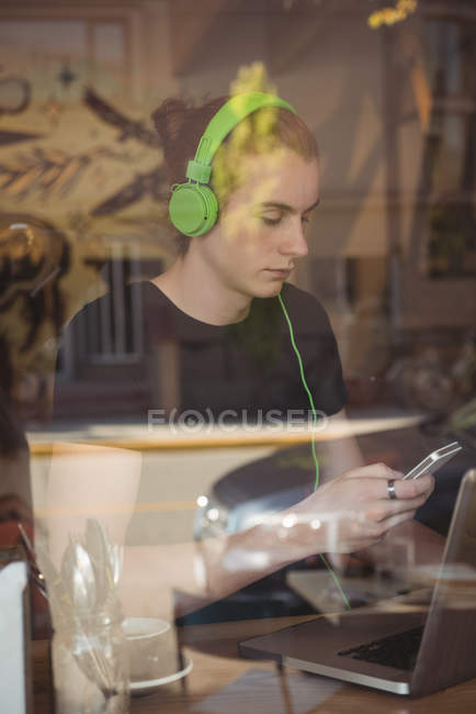 Человек слушает музыку на наушниках во время использования мобильного телефона в кафе — стоковое фото