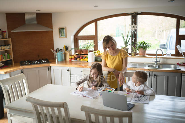 Mãe ajudando crianças com lição de casa na cozinha em casa — Fotografia de Stock