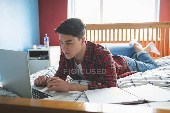 Молодий чоловік працює з ноутбуком, лежачи на ліжку . — стокове фото