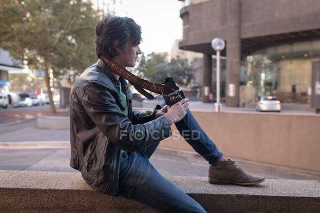 Fotografo con macchina fotografica seduto sul muro vicino alla strada — Foto stock