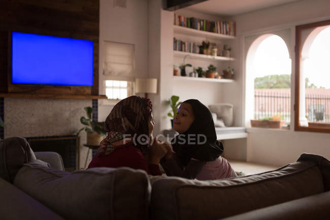 Madre musulmana juguetona e hija tomados de la mano en el sofá en casa - foto de stock