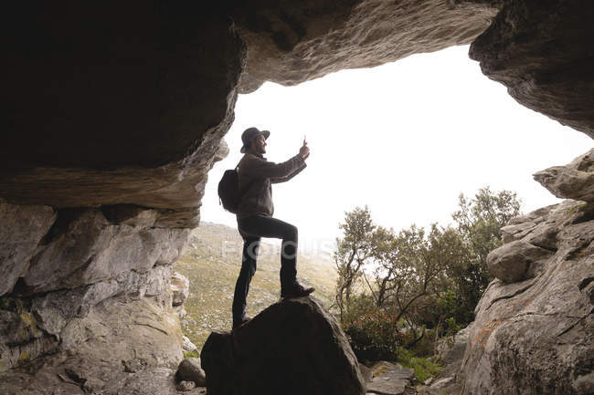 Hiker standing at the cave entrance with backpack taking photos — Stock Photo