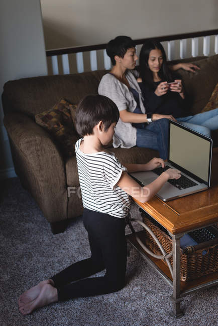 Niño usando el ordenador portátil, mientras que la madre y la hija sentado en segundo plano en casa - foto de stock