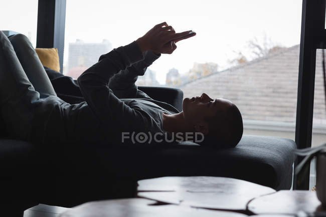 Hombre joven usando el teléfono móvil mientras está acostado en el sofá en la sala de estar en casa - foto de stock