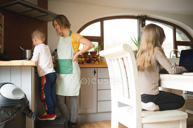 Мати і син готують їжу, поки дочка використовує ноутбук на фоні на кухні вдома — стокове фото