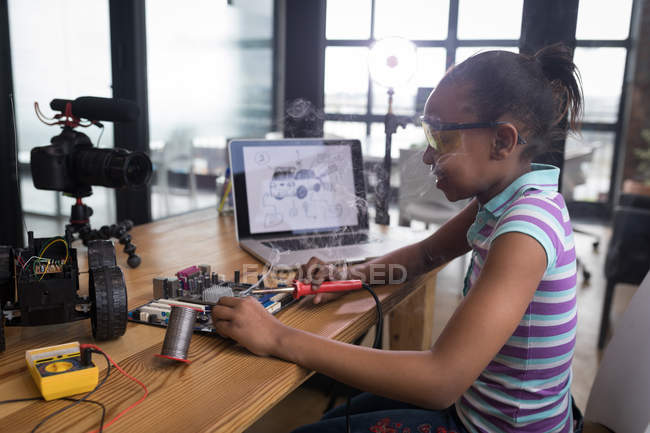 Pre-adolescent blogger girl soldering circuit board of electric toy car in office. — Stock Photo