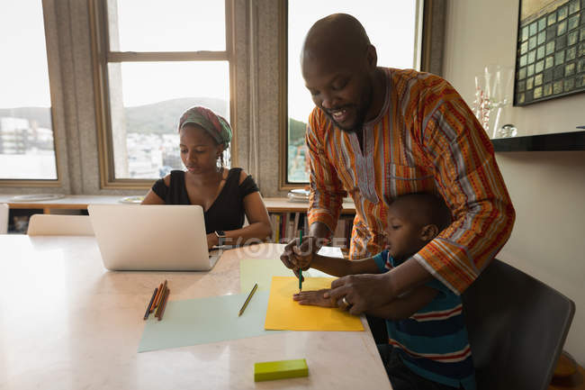 Vater unterstützt Sohn beim Zeichnen von Skizzen am Tisch mit Mutter am heimischen Laptop. — Stockfoto