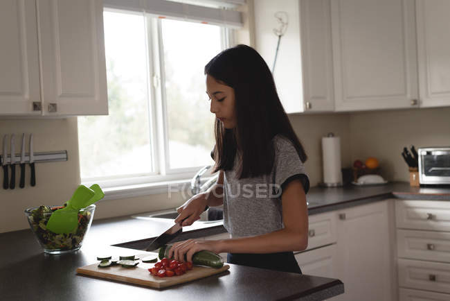 Teenage girl slicing cucumber with knife in kitchen at home — Stock Photo