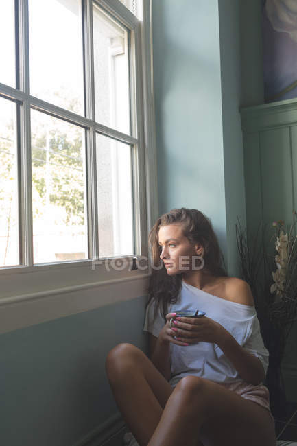 Mujer sentada cerca de la ventana y sosteniendo una taza de café en casa . - foto de stock
