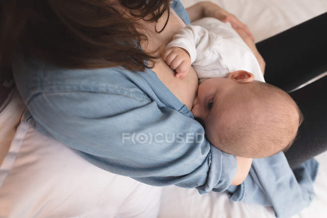 Vue grand angle de la mère assise sur le lit qui allaite son bébé à la maison — Photo de stock