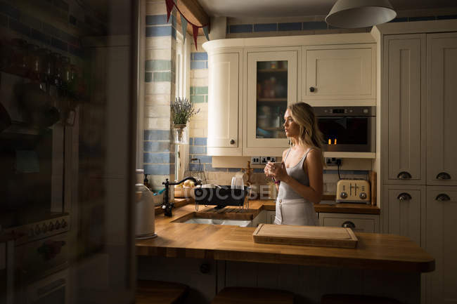 Вид сбоку молодой женщины, стоящей на кухне с кружкой в руках — стоковое фото