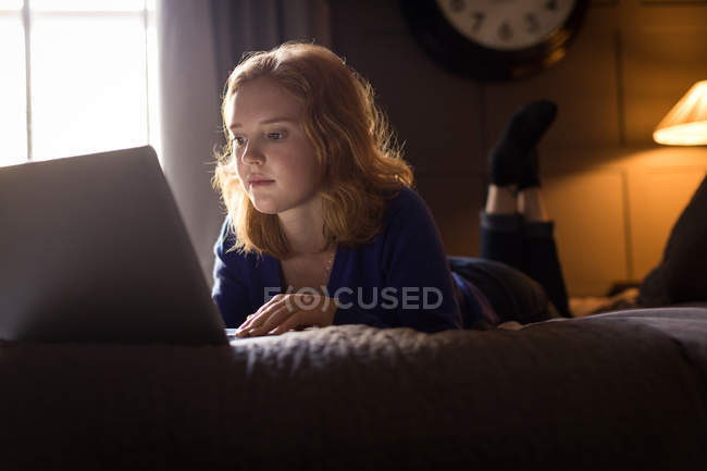Молодая женщина лежит на кровати во время использования ноутбука дома — стоковое фото