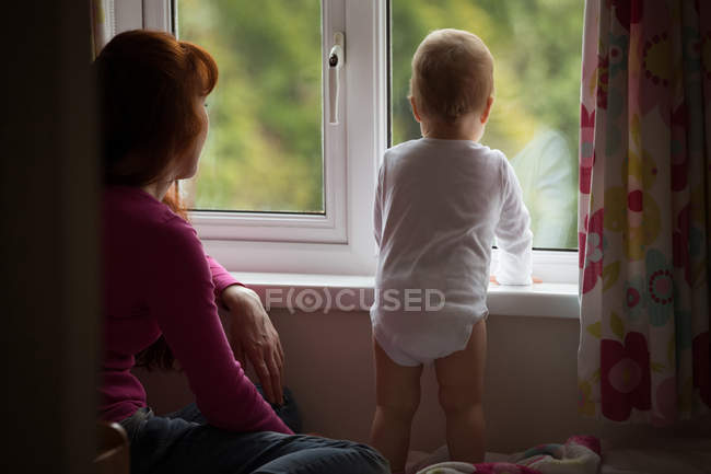 Mother with her baby girl looking through window at home — Stock Photo
