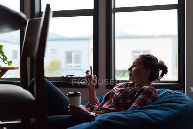 Женщина-руководитель, пользуясь мобильным телефоном, пьет кофе в офисе — стоковое фото