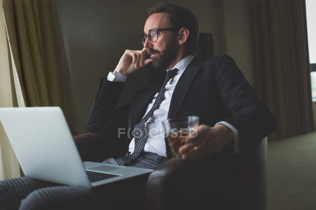 Pensativo hombre de negocios que usa el ordenador portátil mientras toma whisky en la habitación del hotel - foto de stock