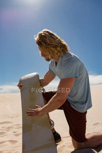 Homme appliquant de la cire de planche de surf sur un plateau de sable au désert par une journée ensoleillée — Photo de stock
