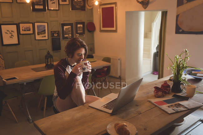 Donna che prende il caffè mentre usa il computer portatile a casa . — Foto stock