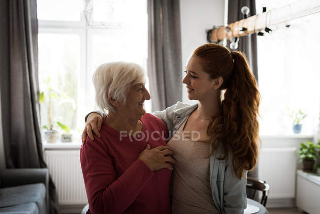 Smiling grandmother and grand daughter standing with arm around in living room — Stock Photo