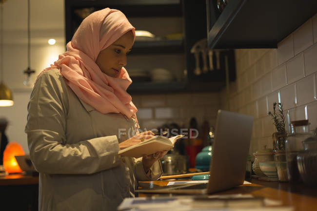 Mulher muçulmana que escreve a receita do computador portátil na cozinha — Fotografia de Stock