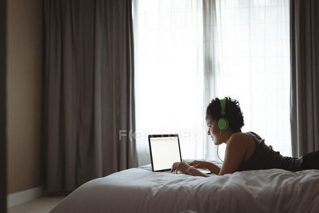 Mulher usando laptop enquanto ouve música na cama no quarto — Fotografia de Stock