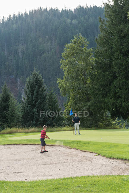 Padre mirando a su hijo mientras juega en el campo de golf en el día soleado - foto de stock