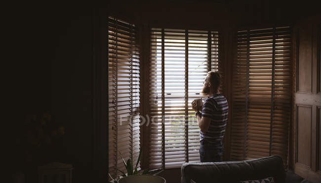 Homme debout près de la fenêtre tout en prenant un café à la maison — Photo de stock