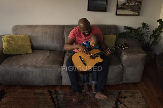 Pai e filho tocando guitarra no sofá na sala de estar em casa . — Fotografia de Stock