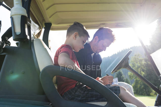 Батько і син сидять у візку для гольфу і пишуть в папері в сонячний день — стокове фото