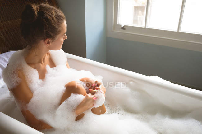 Mujer tomando baño con espuma en la bañera y mirando a través de la ventana en casa . - foto de stock