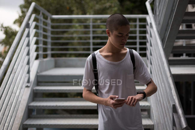 Junger Mann kontrolliert Zeit auf Armbanduhr, während er Handy im Treppenhaus benutzt — Stockfoto