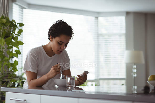 Mujer desayunando mientras usa el teléfono móvil en casa - foto de stock