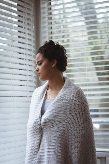 Mujer pensativa envuelta en chal mirando a través de la ventana en casa - foto de stock