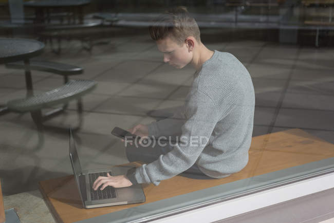 Adolescente niño sosteniendo el teléfono móvil mientras usa el ordenador portátil en la cantina - foto de stock